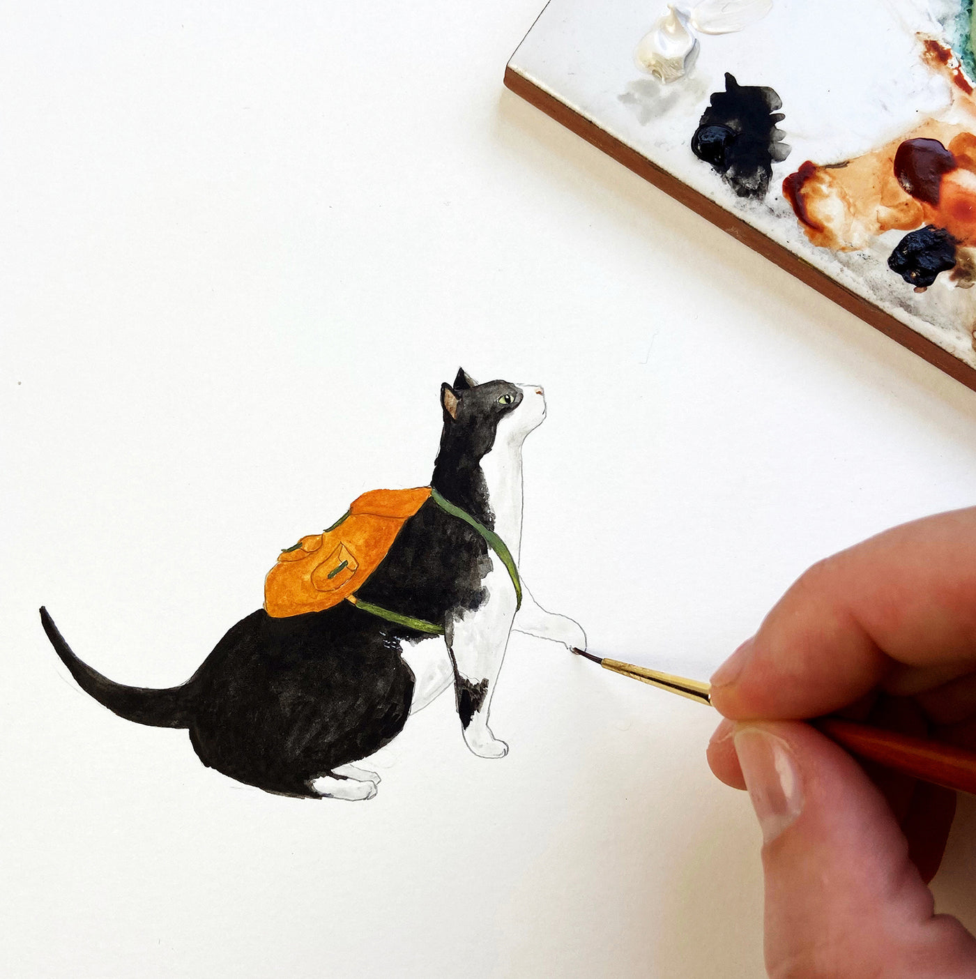 A hand painting a cat with a backpack with watercolour paints